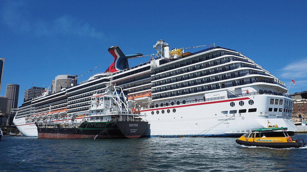 A large cruise ship docked at a passenger terminal on a sunny day with blue sky.