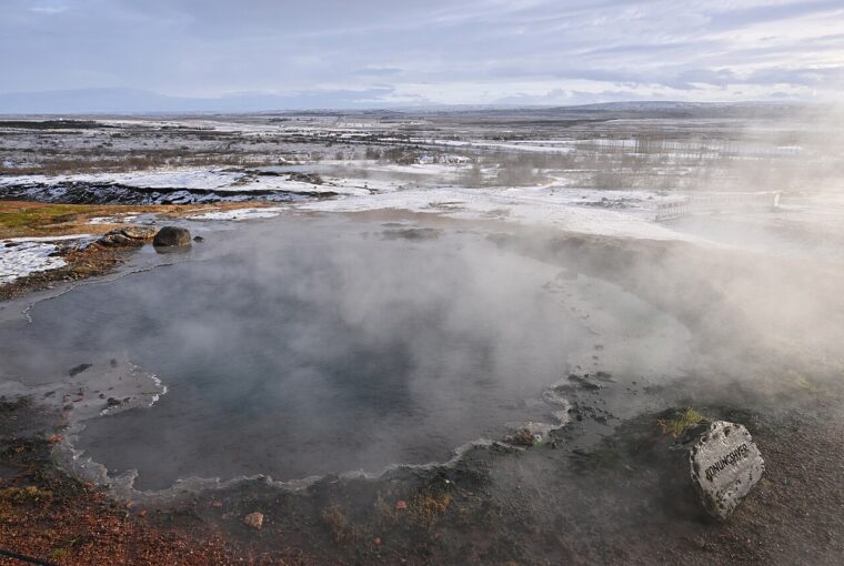 A steaming, natural thermal pool surrounded by a snowy, barren landscape under a cloudy sky in Iceland.