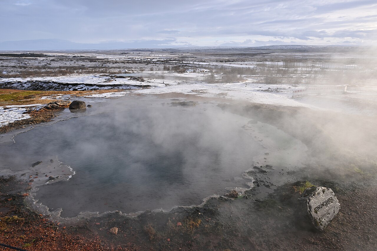A steaming, natural thermal pool surrounded by a snowy, barren landscape under a cloudy sky in Iceland.