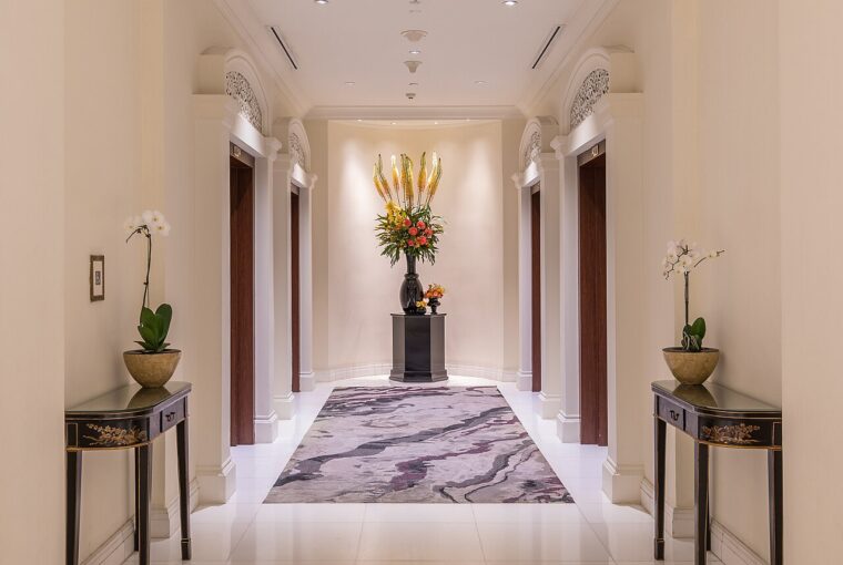 A symmetrical hotel hallway featuring elevators and floral arrangements on console tables, leading to a central bouquet.