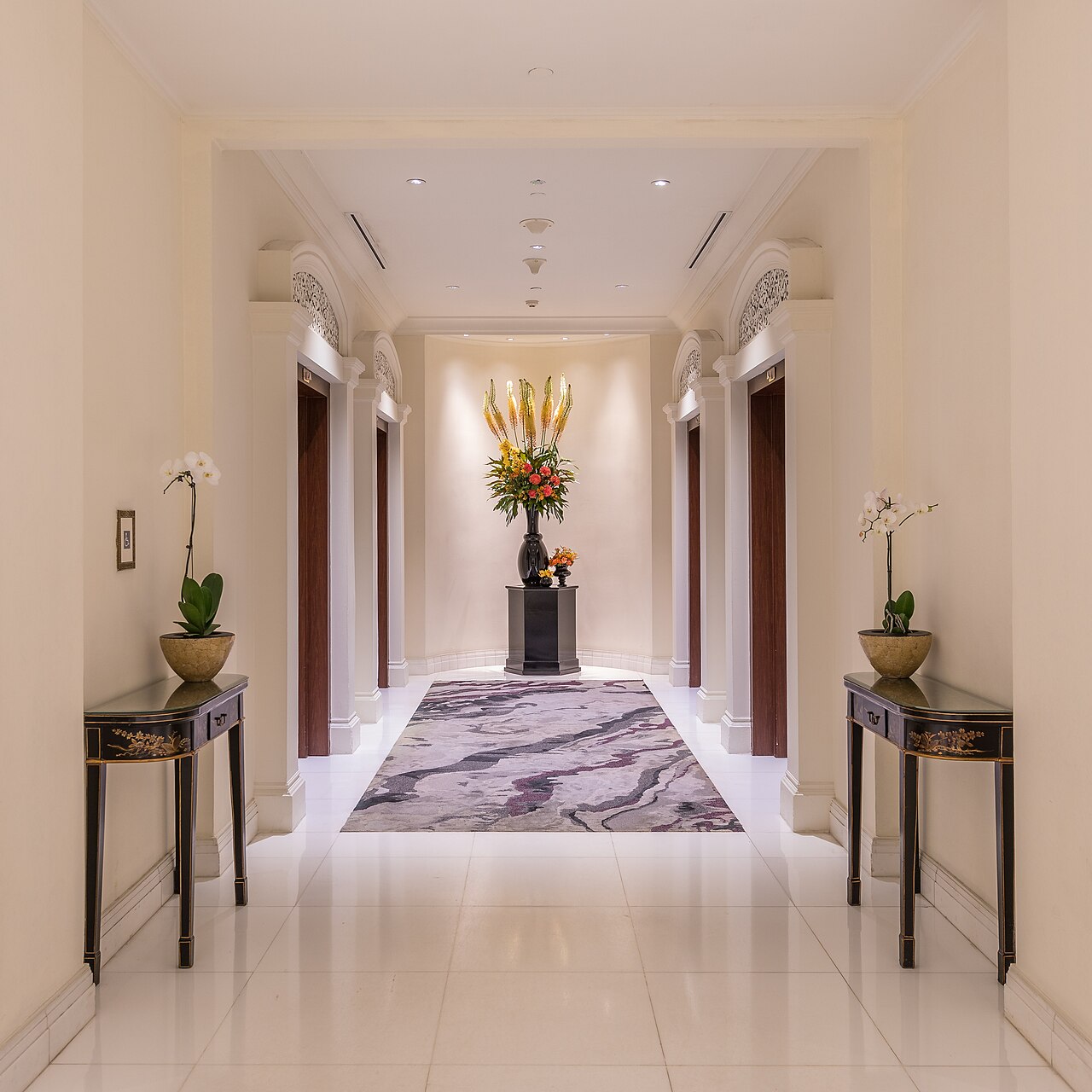 A symmetrical hotel hallway featuring elevators and floral arrangements on console tables, leading to a central bouquet.