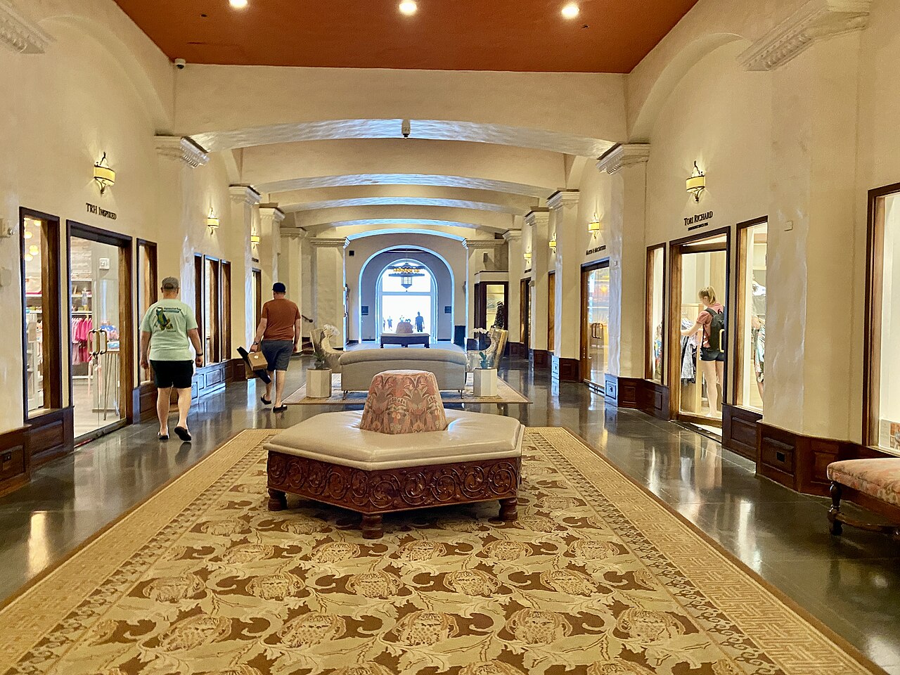 A wide hallway in a historic hotel lobby with polished floors, arched doorways, and guests walking through.