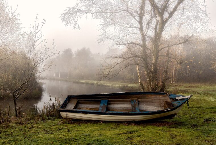 A small wooden boat rests on the grassy bank of a calm, misty lake during an autumn morning.