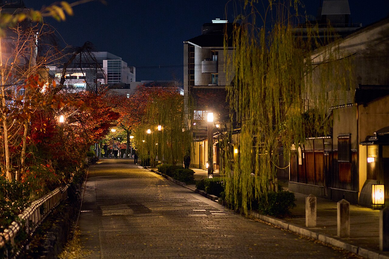 A peaceful, stone-paved street in Kyoto at night, framed by autumn foliage and traditional wooden buildings.