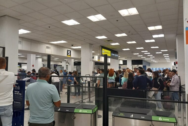 A crowded airport security checkpoint queue with travelers waiting in line near signage and bins.