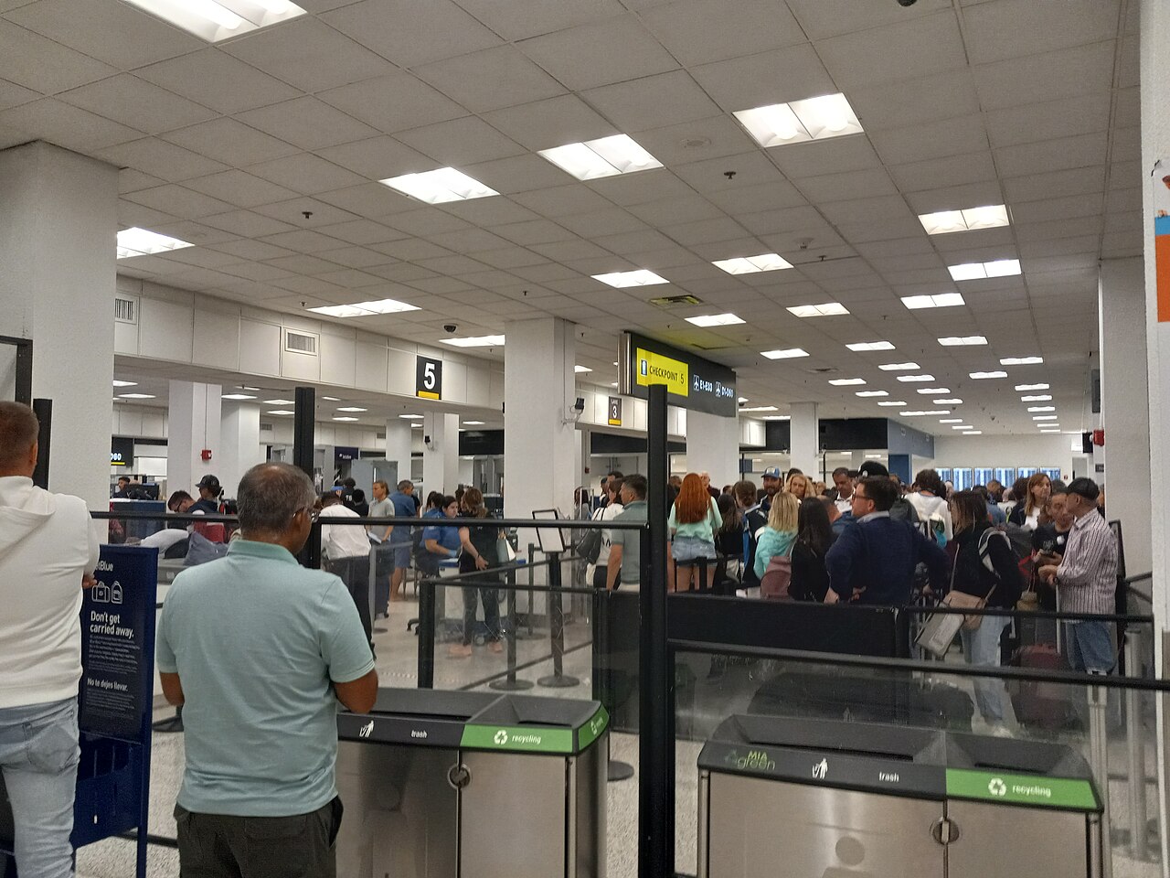A crowded airport security checkpoint queue with travelers waiting in line near signage and bins.