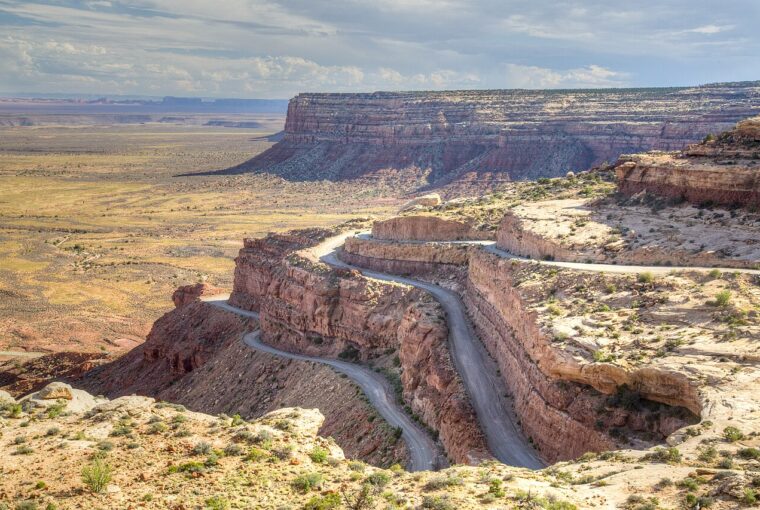 A winding mountain road descends a steep cliffside overlooking a vast, arid desert landscape under a blue sky.