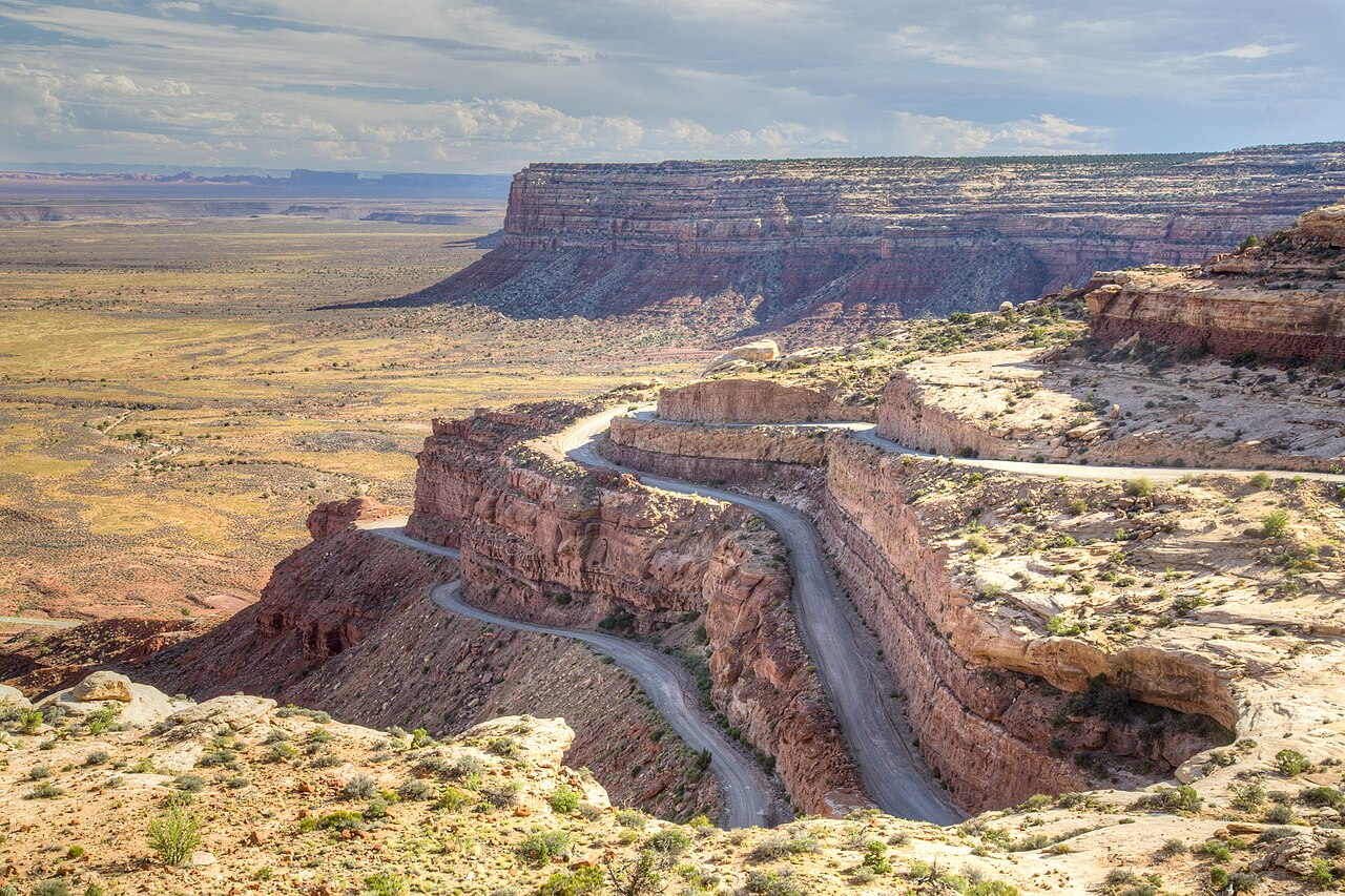 A winding mountain road descends a steep cliffside overlooking a vast, arid desert landscape under a blue sky.